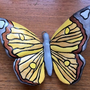 May include: A ceramic butterfly wall decoration with yellow, brown, and grey paint. The butterfly has a detailed design with black lines and a textured surface.