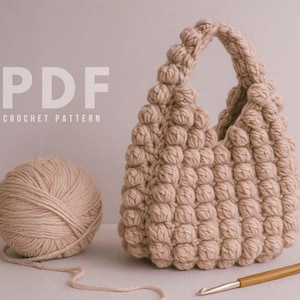 Puede incluir: Un bolso de ganchillo beige con un patrón texturizado en forma de burbujas, una madeja de hilo y un ganchillo. El bolso tiene un asa y está junto a una madeja de hilo. El texto "PDF CROCHET PATTERN" está en la parte superior.