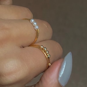 Può includere: Due anelli d'oro su un dito. Un anello ha quattro piccole perle bianche, l'altro ha cinque piccole perle dorate.