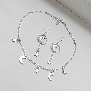 May include: A silver necklace and earrings set with crescent moon and star charms. The necklace has a delicate chain with three crescent moon charms and four star charms. The earrings are dangle earrings with a crescent moon charm and a star charm.
