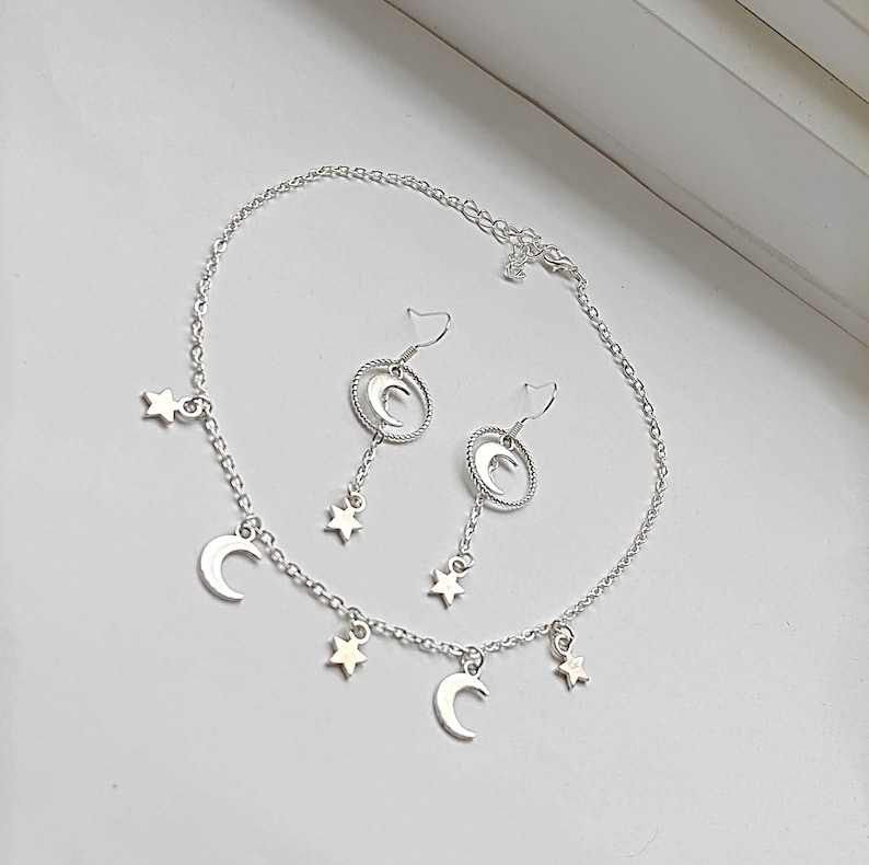 May include: A silver necklace and earrings set with crescent moon and star charms. The necklace has a delicate chain with three crescent moon charms and four star charms. The earrings are dangle earrings with a crescent moon charm and a star charm.