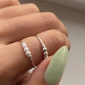 Może przedstawiać: Pokazano dwa srebrne pierścionki. Jeden pierścionek ma rząd małych, białych koralików, a drugi trzy srebrne koraliki. Pierścionki są na palcu z jasnozielonym paznokciem.