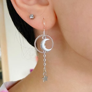 May include: Silver dangle earrings with a crescent moon charm and a star charm hanging from a chain. The moon charm is surrounded by a circle of twisted wire.
