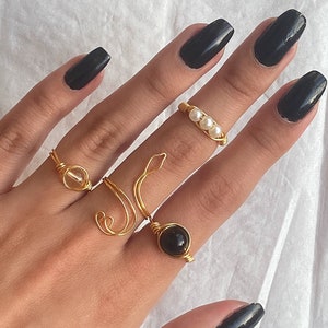 Puede incluir: Una mano con cinco anillos de oro con diferentes dise&ntilde;os. Los anillos presentan una piedra transparente, una piedra negra, un dise&ntilde;o de perlas y dos dise&ntilde;os de alambre simples. La mano est&aacute; pintada con esmalte de u&ntilde;as negro.