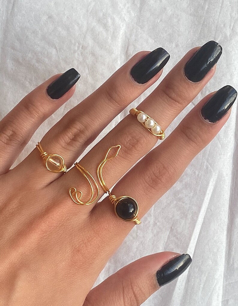 Puede incluir: Una mano con cinco anillos de oro con diferentes dise&ntilde;os. Los anillos presentan una piedra transparente, una piedra negra, un dise&ntilde;o de perlas y dos dise&ntilde;os de alambre simples. La mano est&aacute; pintada con esmalte de u&ntilde;as negro.