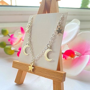 May include: A silver chain necklace with three crescent moon charms and three star charms. The necklace is displayed on a wooden easel.