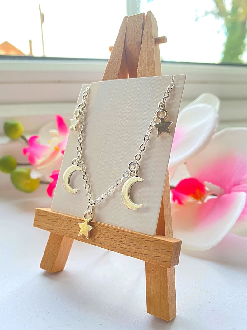 May include: A silver chain necklace with three crescent moon charms and three star charms. The necklace is displayed on a wooden easel.