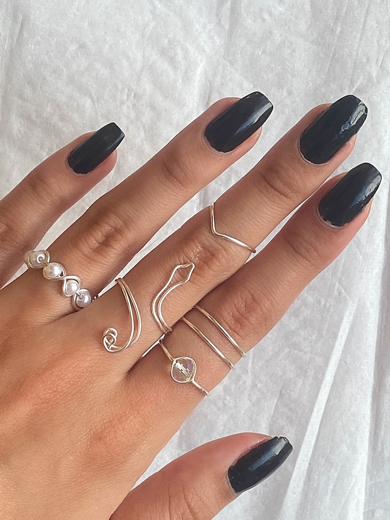Puede incluir: Una mano con esmalte de u&ntilde;as negro lleva un conjunto de anillos de plata. Los anillos incluyen un anillo de perlas, un anillo de serpiente, un anillo de cristal y tres bandas finas.