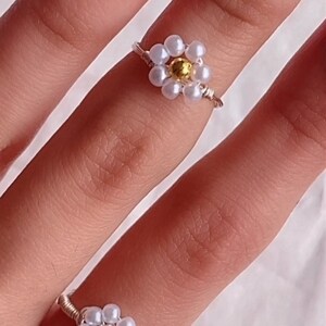 Puede incluir: Dos anillos de plata con dise&ntilde;os de flores de perlas blancas. El anillo m&aacute;s grande tiene una cuenta central dorada. Los anillos se llevan en un dedo con esmalte de u&ntilde;as rojo.