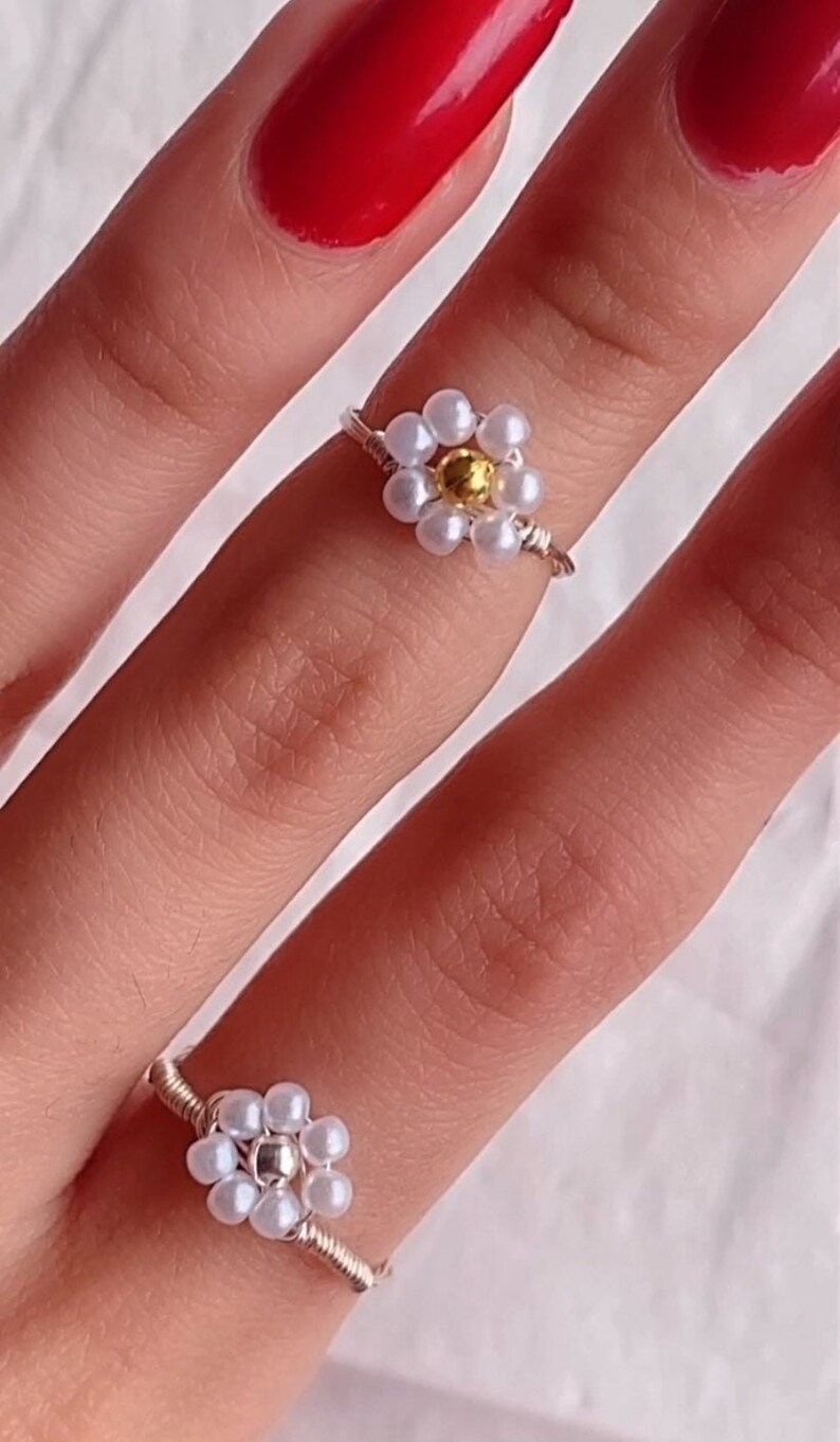 Puede incluir: Dos anillos de plata con dise&ntilde;os de flores de perlas blancas. El anillo m&aacute;s grande tiene una cuenta central dorada. Los anillos se llevan en un dedo con esmalte de u&ntilde;as rojo.
