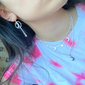 May include: A silver necklace with a crescent moon charm and star charms. The necklace is layered with a second chain with a crescent moon charm. The necklace is worn by a person with long dark hair. The person is wearing a pink and white tie-dye shirt.