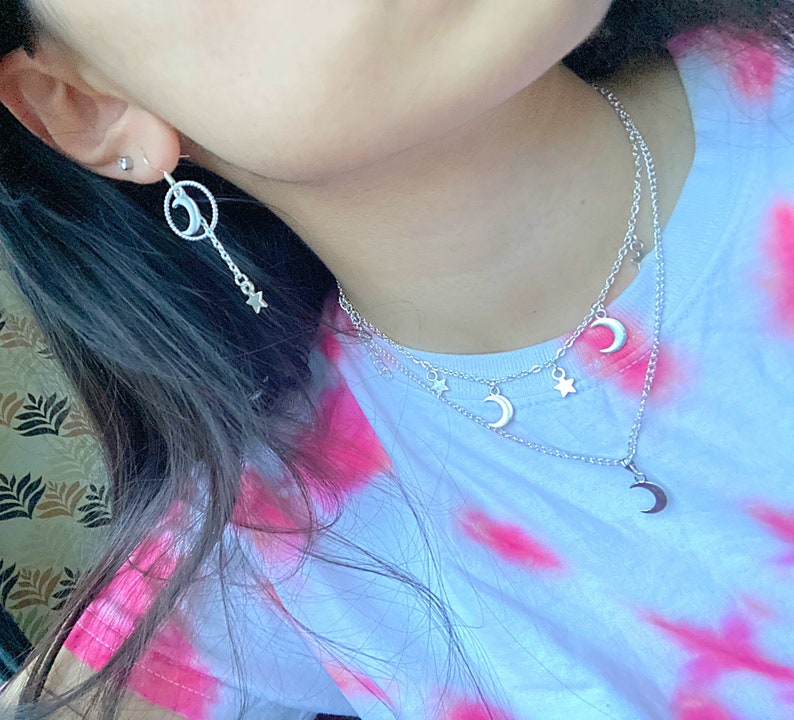 May include: A silver necklace with a crescent moon charm and star charms. The necklace is layered with a second chain with a crescent moon charm. The necklace is worn by a person with long dark hair. The person is wearing a pink and white tie-dye shirt.