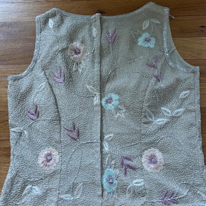 May include: A sleeveless, beige top with floral embroidery. The top features embroidered flowers in shades of pink and blue, with purple leaves and white vines. The fabric has a textured pattern and is trimmed with small beads.