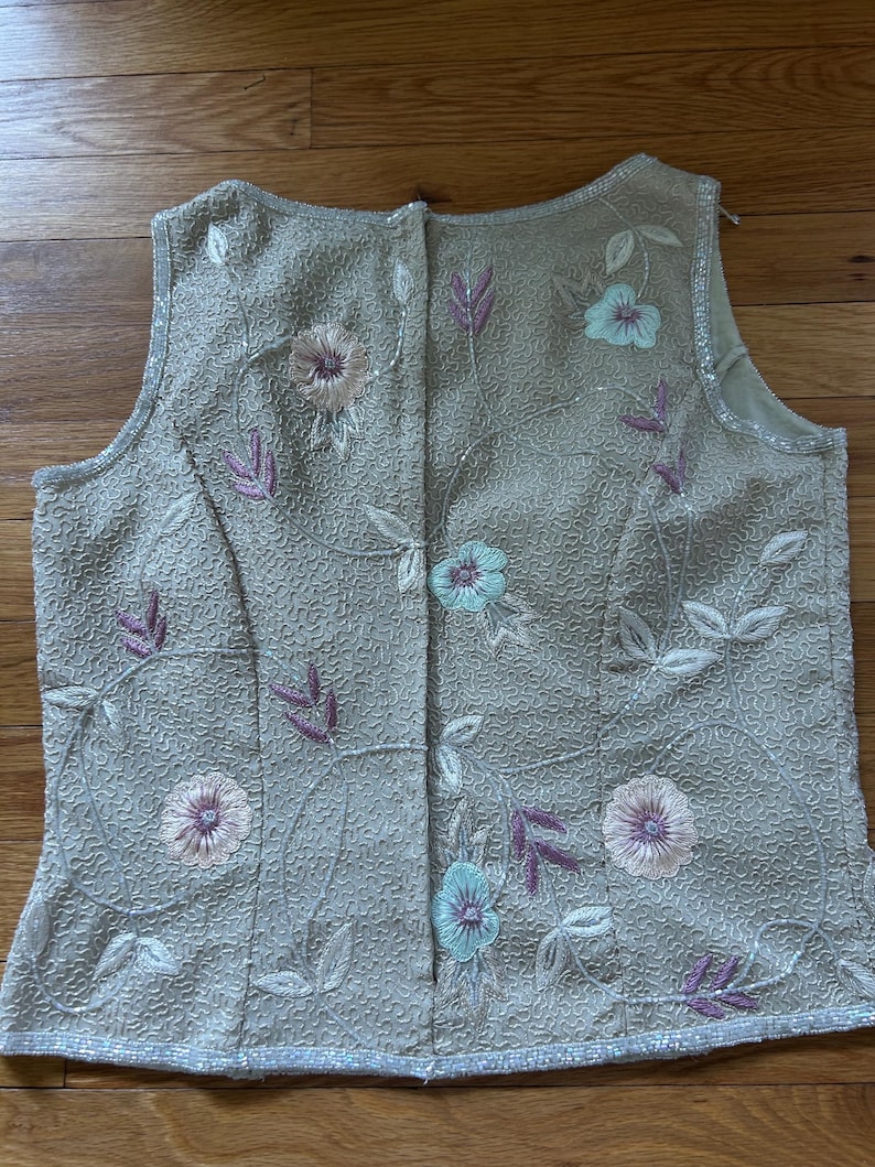May include: A sleeveless, beige top with floral embroidery. The top features embroidered flowers in shades of pink and blue, with purple leaves and white vines. The fabric has a textured pattern and is trimmed with small beads.