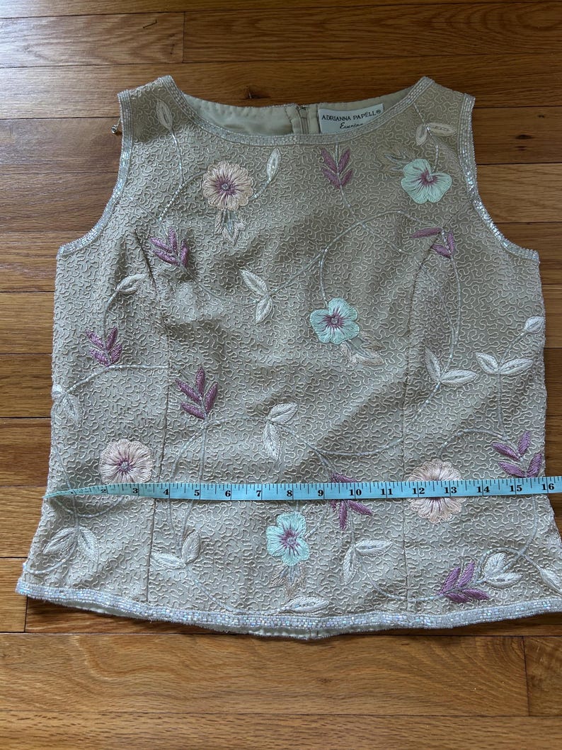 May include: Beige sleeveless top with floral embroidery in pink, blue, and white. The top features a textured fabric and a zipper closure in the back. The waist measures approximately 16 inches.