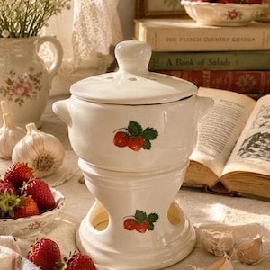 May include: A white ceramic garlic baker with a lid, featuring a strawberry design. The baker is stacked on a base with openings. Fresh strawberries, garlic, and a floral bowl are nearby, suggesting a kitchen or dining theme.