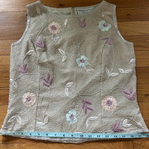 May include: Beige sleeveless top with floral embroidery in pink, lavender, and light blue. The top features a textured fabric and a beaded neckline. The brand name "ADRIANNA PAPELL" is visible on the top. A measuring tape is placed across the bottom of the top.