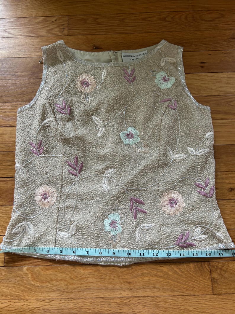 May include: Beige sleeveless top with floral embroidery in pink, lavender, and light blue. The top features a textured fabric and a beaded neckline. The brand name "ADRIANNA PAPELL" is visible on the top. A measuring tape is placed across the bottom of the top.