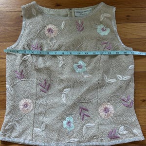 May include: Beige sleeveless top with floral embroidery. The top features a round neckline and is embellished with sequins. The floral embroidery includes pink, blue, and purple flowers and leaves. The top measures approximately 16 inches across.