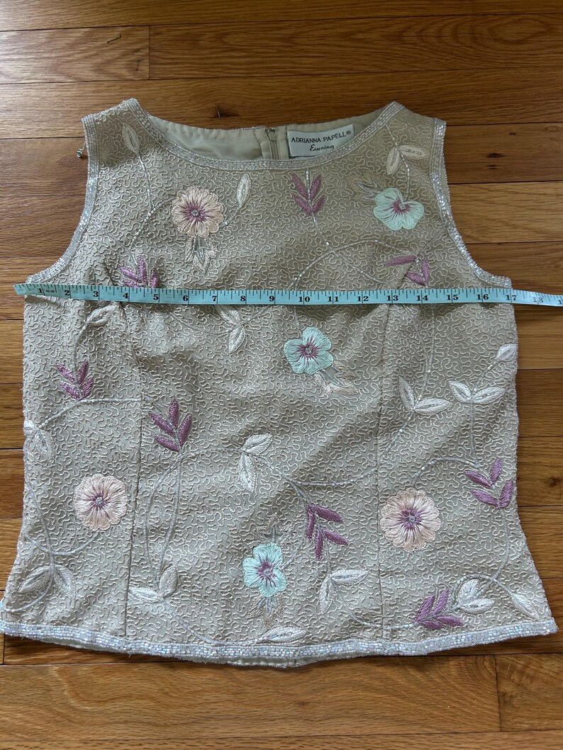 May include: Beige sleeveless top with floral embroidery. The top features a round neckline and is embellished with sequins. The floral embroidery includes pink, blue, and purple flowers and leaves. The top measures approximately 16 inches across.