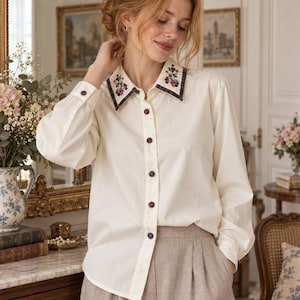 Puede incluir: Una camisa de manga larga color crema con cuello en punta que presenta bordados florales oscuros y un borde a cuadros. La camisa tiene una tapeta frontal con botones circulares oscuros y mangas largas con puños abotonados, junto a pantalones beige.