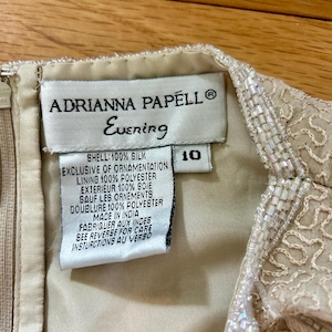 May include: Close-up of an Adrianna Papell Evening dress tag. The tag is cream-colored with black text, including the brand name, "Evening," and size 10. The dress is made of silk and polyester, with beaded embellishments.