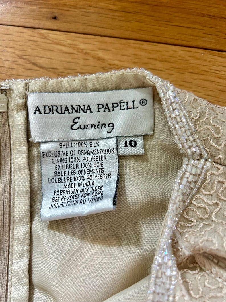May include: Close-up of an Adrianna Papell Evening dress tag. The tag is cream-colored with black text, including the brand name, "Evening," and size 10. The dress is made of silk and polyester, with beaded embellishments.