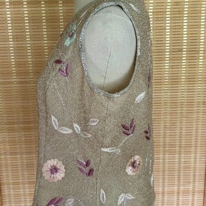 May include: A sleeveless, tan-colored top adorned with floral and leaf embroidery in shades of pink, purple, and green. The top is embellished with sequins and has a beaded trim along the neckline and hem.