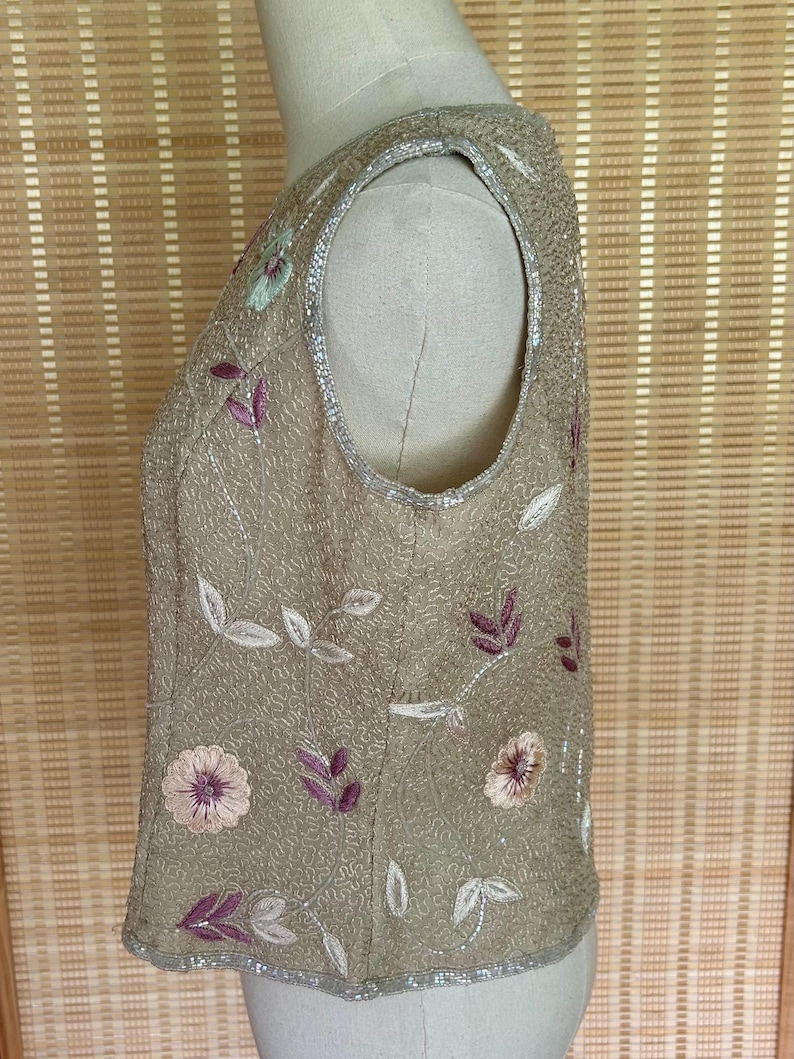May include: A sleeveless, tan-colored top adorned with floral and leaf embroidery in shades of pink, purple, and green. The top is embellished with sequins and has a beaded trim along the neckline and hem.