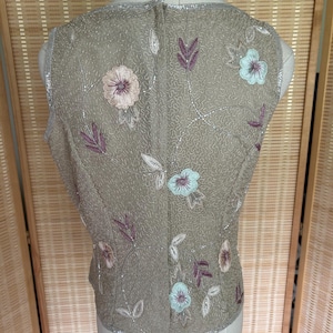 May include: A sleeveless, beaded vest in a muted olive green hue. The vest is adorned with embroidered floral and leaf patterns in shades of pink, blue, and purple. Silver beading trims the edges, adding a touch of sparkle. The vest is displayed on a mannequin.