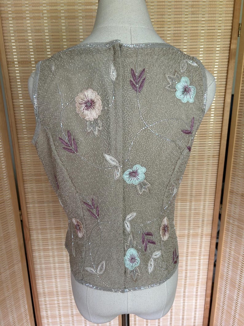May include: A sleeveless, beaded vest in a muted olive green hue. The vest is adorned with embroidered floral and leaf patterns in shades of pink, blue, and purple. Silver beading trims the edges, adding a touch of sparkle. The vest is displayed on a mannequin.