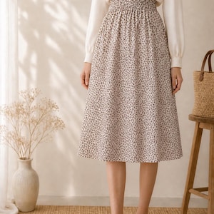 May include: A woman's outfit featuring a white long-sleeve blouse and a knee-length skirt with a small, dark pattern on a light background. The outfit is paired with beige flats. The background includes a woven rug and a wooden stool.