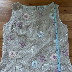 May include: A sleeveless, beige beaded top with floral embroidery in pink, purple, and light blue. The top features a round neckline and a beaded hemline. The brand name "ADRIANNA PAPELL" is visible on the label. A measuring tape is placed next to the top.