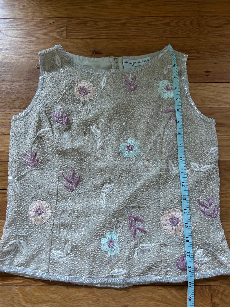 May include: A sleeveless, beige beaded top with floral embroidery in pink, purple, and light blue. The top features a round neckline and a beaded hemline. The brand name "ADRIANNA PAPELL" is visible on the label. A measuring tape is placed next to the top.