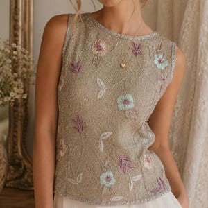 May include: A sleeveless, beaded top in a champagne color, adorned with embroidered floral and leaf patterns in pastel shades of pink, purple, and blue. The top has a silver trim around the neckline and armholes, and a small gold heart pendant.