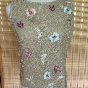 May include: A sleeveless, tan-colored top with floral embroidery. The top features embroidered flowers in shades of pink and blue, along with purple leaves and silver beading around the neckline and hem. The fabric has a textured, patterned appearance.