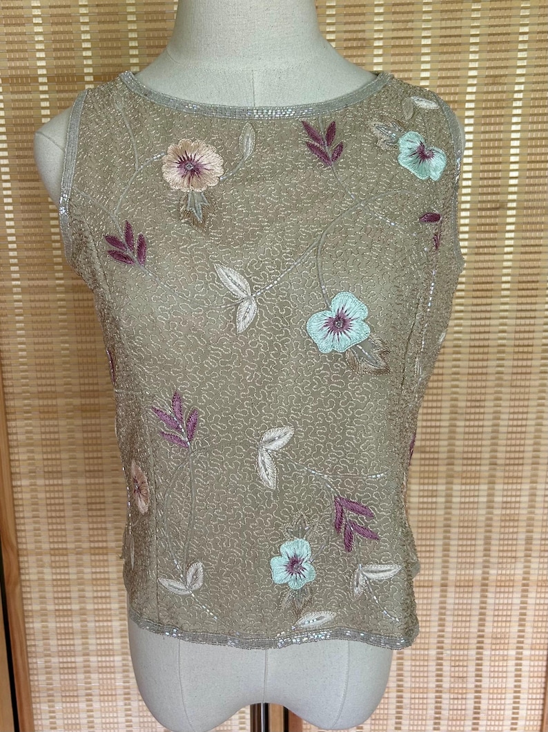 May include: A sleeveless, tan-colored top with floral embroidery. The top features embroidered flowers in shades of pink and blue, along with purple leaves and silver beading around the neckline and hem. The fabric has a textured, patterned appearance.