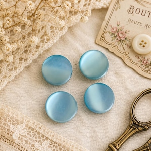 May include: Four round, light blue buttons with a pearlescent sheen are arranged on a cream-coloured fabric. The buttons are surrounded by vintage sewing notions, including scissors, a spool of thread, and a card that reads "BOUTONS".