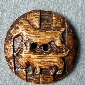 May include: A round, dark brown button featuring a carved, raised relief design of a goat standing in profile. The button has a textured, wood-like appearance with two central holes for attachment, showcasing a detailed, rustic, and natural aesthetic.