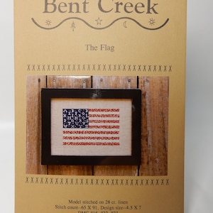 May include: A cross-stitch pattern chart titled The Flag by Bent Creek on tan paper. It features a framed image of a flag with an alphabet in the blue field and red and white stripes. Text includes stitch count 65 X 91 and design size 4.5 X 7 inches.