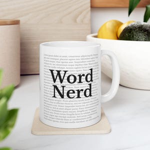 May include: White ceramic mug with the words "Word Nerd" in a bold, black font. The mug is surrounded by a background of blurred text. The mug sits on a beige coaster.