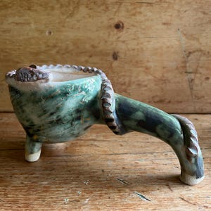 May include: A ceramic vessel with a mottled green and brown glaze, featuring a wide bowl supported by three short legs and a long, curved handle extending from the side. The rim and handle are decorated with a textured, notched edge pattern.