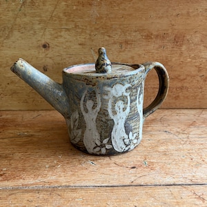 May include: A handcrafted ceramic teapot with a lid and handle. The teapot features white figures, flowers, and leaves on a brown and blue surface. The spout is long and angled, and the handle is curved.