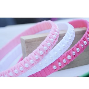 Könnte beinhalten: Drei Haarbänder in Pink, Weiß und Hellrosa, jedes mit weißen Perlenakzenten. Die Haarbänder sind geflochten und haben eine Satin-Oberfläche.