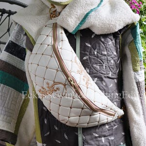 Könnte beinhalten: Gesteppte Patchwork-Jacke mit weißer, fleecegefütterter Kapuze und weißer, gesteppter Bauchtasche. Die Bauchtasche hat einen goldenen Reißverschluss und ein goldgesticktes Design. Die Jacke ist mehrfarbig mit grünen, grauen und weißen Einsätzen.