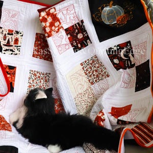 Op de afbeelding: Een handgemaakte Halloween-quilt met een patchwork-ontwerp. De quilt heeft witte vierkanten met oranje, zwarte en rode geborduurde patronen van pompoenen, katten en andere feestelijke motieven. Een zwart-witte kat rust op de quilt.