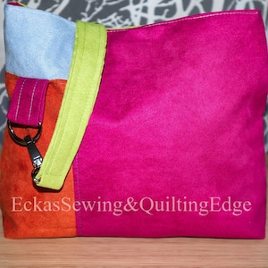 Può includere: Una borsa a tracolla patchwork colorata con un cinturino verde lime. La borsa è realizzata in un materiale morbido, simile al camoscio, e presenta un design rosa acceso, arancione e blu chiaro. La borsa ha una chiusura argentata e il testo "Eckas Sewing & Quilting Edge" è stampato sul davanti.