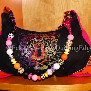 Puede incluir: Bolso de mano negro con un diseño bordado de cara de leopardo. El bolso presenta ribetes rosas, detalles en naranja y una correa de cuentas con cuentas de colores. El texto "Eckas Sewing & Quiting Edge" es visible en el bolso.