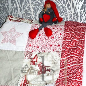 Peut inclure: Une courtepointe en patchwork présentant une variété de motifs en tissu rouge et blanc, incluant des scènes d'hiver avec des maisons, des rennes et des motifs floraux. Une poupée en peluche avec un bonnet rouge et un cœur sur la poitrine.