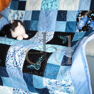 May include: A patchwork quilt featuring various shades of blue, including navy, sky blue, and teal, with floral patterns, solid squares, and embroidered cat motifs. A black and white plush cat toy sits on the quilt, which has the text eckshomedesignedge.